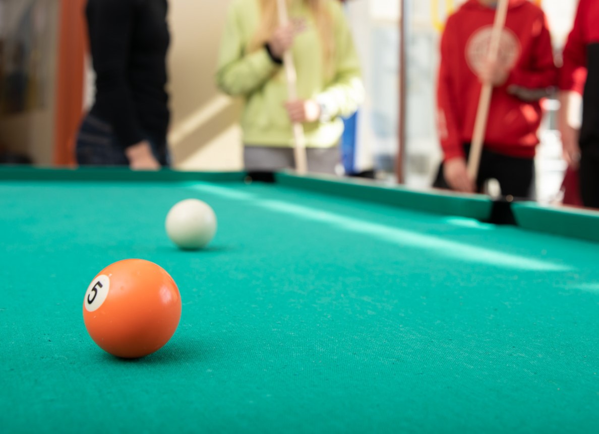 Oranssi biljardipallo biljardipöydällä, taustalla neljä nuorta.